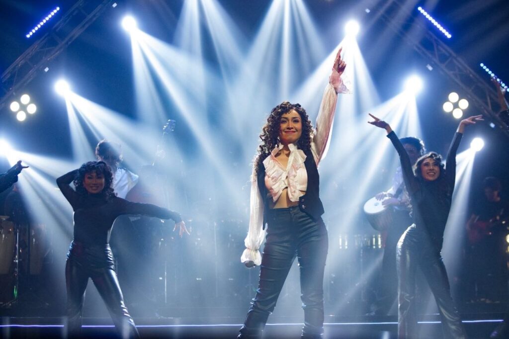 Molly Langley stands center stage as Gloria Estefan, raising one arm beneath bright concert lights while dancers perform around her in On Your Feet!.
